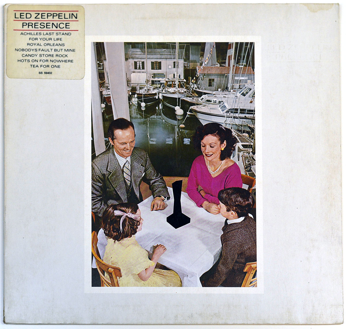 Album cover of Led Zeppelin's 'Presence' featuring a vintage-style family scene with a mysterious black object at the center of a table, surrounded by four people smiling. A marina with boats and reflections in the water serves as the background. The album title and tracklist appear in a label at the top left corner.